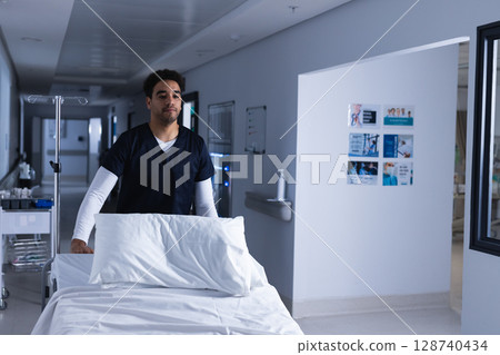 Young man pushing empty hospital bed down bright hospital corridor, with IV pole and supply trolley Young man pushing empty hospital bed down bright hospital corridor, with IV pole and supply trolley 128740434