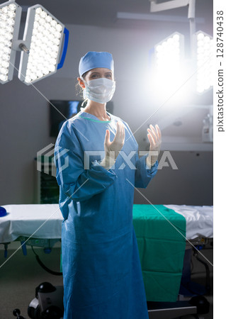 Female surgeon standing in operating room under overhead LED lights beside green draped table 128740438