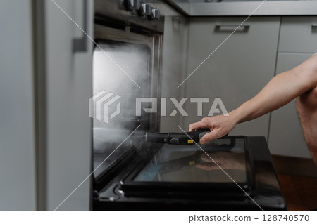 Side view of cleaning service employee male cleaning oven with steam cleaner in kitchen, holding steam cleaner gun removing grease and ensuring hygienic cooking environment, close-up cropped shot 128740570