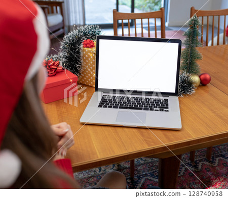 Woman using laptop in cozy dining room, with holiday gift boxes wreath lights and snow-dusted tree 128740958