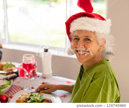 Senior woman sitting in dining room wearing Santa hat, enjoying meal with confetti, copy space Senior woman sitting in dining room wearing Santa hat, enjoying meal with confetti, copy space 128740960
