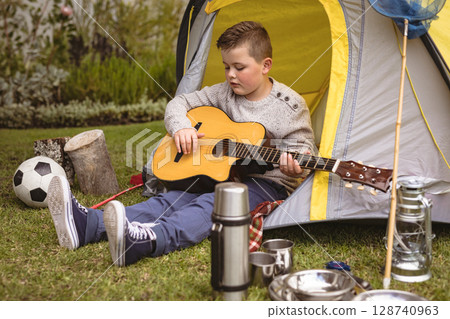 School-aged boy sitting on backyard lawn before yellow camping tent, playing acoustic guitar 128740963