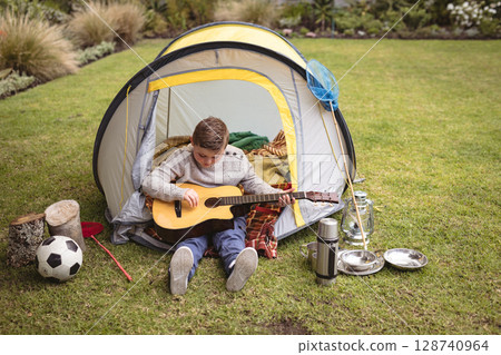 Child boy sitting at backyard tent, strumming guitar with soccer ball and butterfly net nearby 128740964
