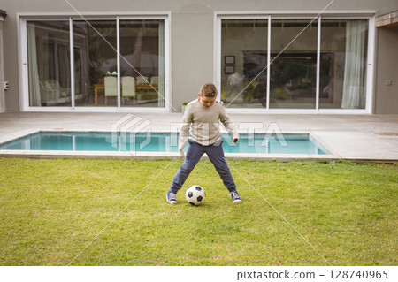 young boy dribbling soccer ball on backyard lawn in front of modern house, featuring pool young boy dribbling soccer ball on backyard lawn in front of modern house, featuring pool 128740965