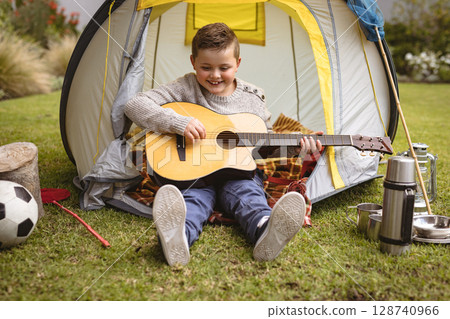 child boy strumming acoustic guitar in backyard, with yellow-gray tent and camping gear 128740966
