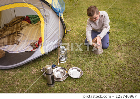 Child boy kneeling next to yellow-trim tent on grassy lawn handling log, with lantern and thermos 128740967
