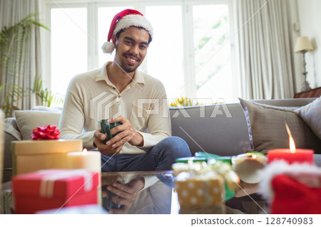 Man wearing Santa hat drinking from green mug in living room, with wrapped gifts and candles 128740983
