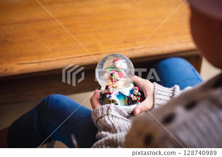 Child holding snow globe with snowman figurine and festive figurines indoors on wooden table 128740989