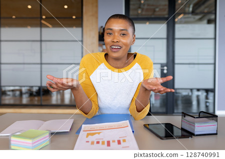 African American woman gesturing with hands at desk in modern office, using tablet and chart 128740991