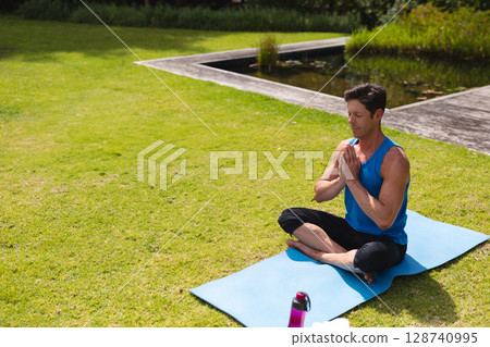 Man practicing yoga on blue yoga mat at garden pond, with pink water bottle, copy space 128740995