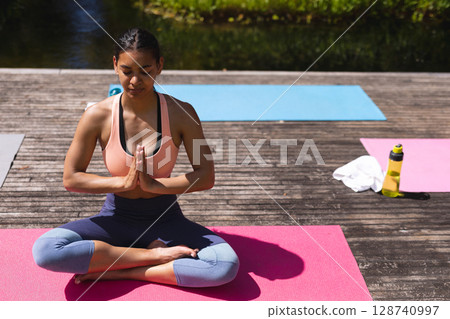 Woman meditating cross-legged on pink yoga mat on deck by pond, with towel and yellow bottle 128740997