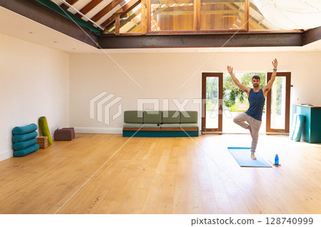 Male practicing tree pose on blue mat in yoga room, with bolsters, water bottle, copy space Male practicing tree pose on blue mat in yoga room, with bolsters, water bottle, copy space 128740999