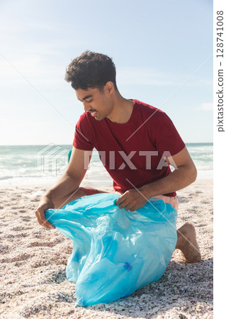 Man kneeling on sandy beach at shoreline, collecting litter with large light blue plastic bag Man kneeling on sandy beach at shoreline, collecting litter with large light blue plastic bag 128741008