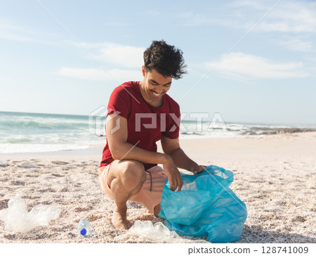 Man kneeling on beach at water edge, collecting plastic debris and water bottle into blue bag Man kneeling on beach at water edge, collecting plastic debris and water bottle into blue bag 128741009