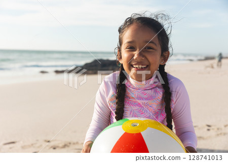 Child girl holding colorful beach ball and smiling while standing on sandy beach by water edge Child girl holding colorful beach ball and smiling while standing on sandy beach by water edge 128741013