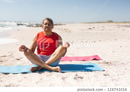 Senior man practicing seated meditation on blue yoga mat at sandy beach with red yoga mat 128741016