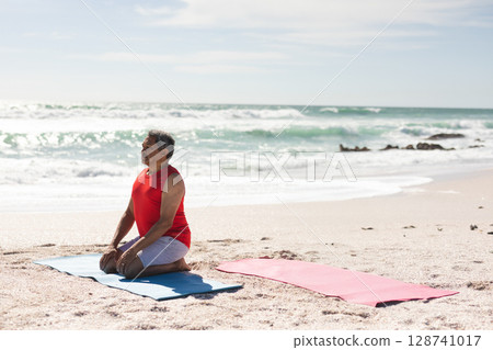 Senior man kneeling on blue yoga mat at sandy beach near shoreline rocks, copy space 128741017