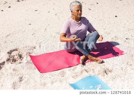 Senior woman practicing yoga on red and blue mats on sandy beach with shells, copy space 128741018
