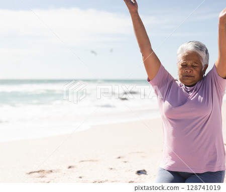 Senior woman stretching arms overhead on sandy beach, with birds flying over calm ocean, copy space 128741019