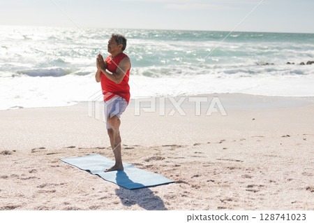 Senior man performing tree pose on beach by ocean, using light blue yoga mat, copy space Senior man performing tree pose on beach by ocean, using light blue yoga mat, copy space 128741023