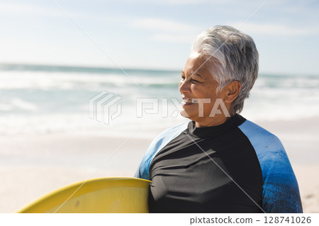 Senior woman standing with yellow surfboard on beach in black or blue wetsuit, enjoying ocean breeze Senior woman standing with yellow surfboard on beach in black or blue wetsuit, enjoying ocean breeze 128741026