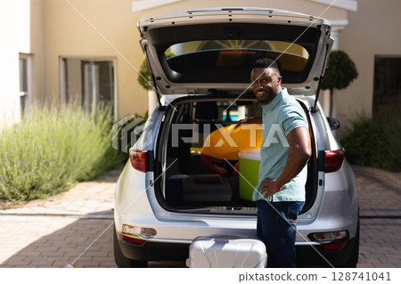 African American man standing by silver SUV trunk in driveway, with cooler and suitcase, copy space 128741041