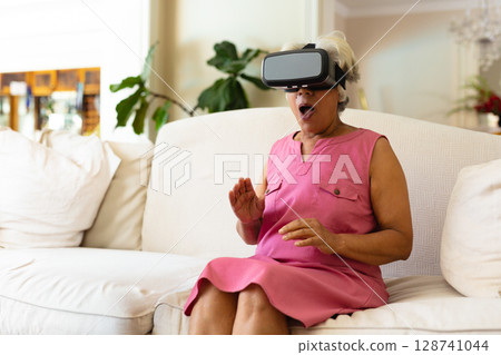 Senior African American woman reacting with surprise wearing VR headset on sofa in living room 128741044