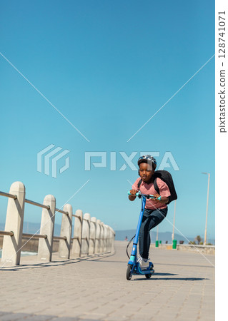 African American child riding e-scooter along seaside promenade, with helmet, backpack, copy space 128741071