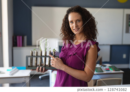 Woman standing facing camera in modern science lab, pointing at test tube rack with seedlings Woman standing facing camera in modern science lab, pointing at test tube rack with seedlings 128741082