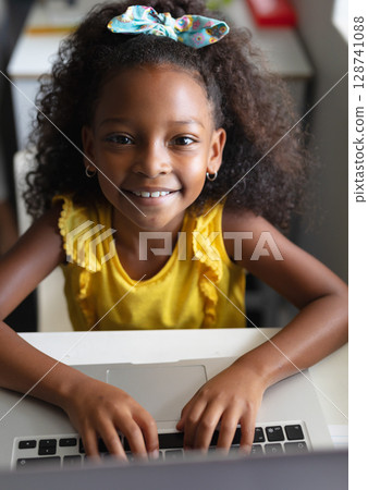 Young girl typing on silver laptop at home study desk, wearing colorful hair bow and smiling 128741088