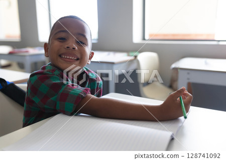 Boy sitting at school desk with open notebook in bright classroom, holding green pencil, copy space 128741092