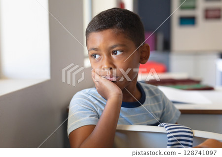7-year-old boy resting cheek on hand and gazing outside at classroom desk by window, with papers 128741097