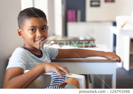 Young boy sitting turned around in chair at classroom desk, holding backpack strap and textbooks Young boy sitting turned around in chair at classroom desk, holding backpack strap and textbooks 128741098