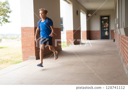 School-aged boy running along covered school corridor with blue backpack and flip-flops, copy space 128741112