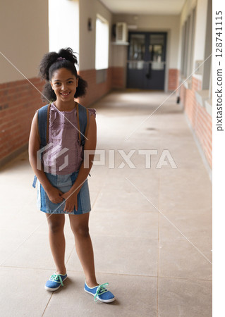 Schoolgirl standing in school corridor near windows, holding blue backpack and wearing sneakers Schoolgirl standing in school corridor near windows, holding blue backpack and wearing sneakers 128741115