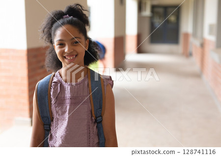 Female child student standing in school walkway with brick columns, windows, wearing blue backpack 128741116