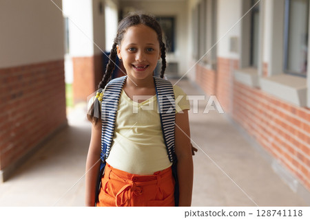 Girl child standing and smiling in school corridor, wearing striped backpack and yellow hair clips Girl child standing and smiling in school corridor, wearing striped backpack and yellow hair clips 128741118