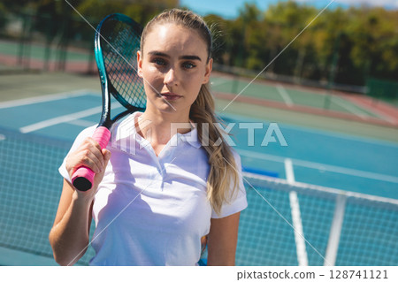 Young woman standing at net on outdoor tennis court, holding racket with pink grip 128741121