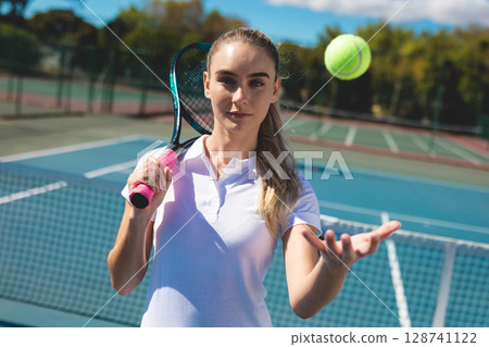 Female tennis player holding racket with pink grip, tossing ball near net on court, preparing serve 128741122
