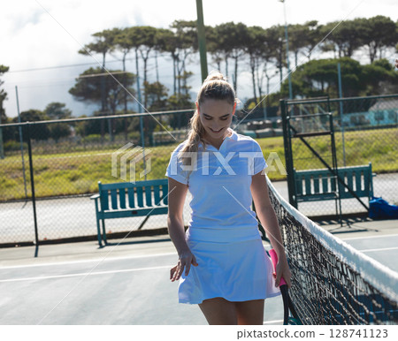 Woman walking alongside tennis net on outdoor tennis court, holding tennis racket Woman walking alongside tennis net on outdoor tennis court, holding tennis racket 128741123