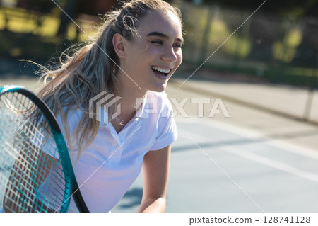 Woman laughing and leaning on outdoor tennis court, holding teal racket near chain-link fence Woman laughing and leaning on outdoor tennis court, holding teal racket near chain-link fence 128741128