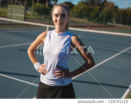Young adult tennis player standing with hands on hips on hard court, showing net, wristband 128741129