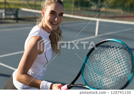 Woman tennis player holding racket on outdoor tennis court smiling at camera, showing wristband 128741130