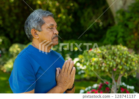 Senior man practicing prayer gesture in private garden, wearing wristwatch and blue t-shirt 128741137