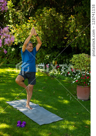 Senior man practicing yoga tree pose in backyard, with gray mat, purple dumbbells, terra-cotta pot 128741138