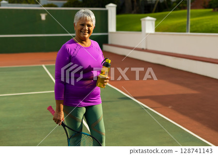 Senior woman standing on tennis court holding racket and yellow water bottle under sunny sky Senior woman standing on tennis court holding racket and yellow water bottle under sunny sky 128741143