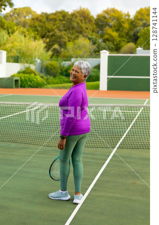 Senior woman practicing on outdoor tennis court, holding tennis racket and smiling by net and lines Senior woman practicing on outdoor tennis court, holding tennis racket and smiling by net and lines 128741144