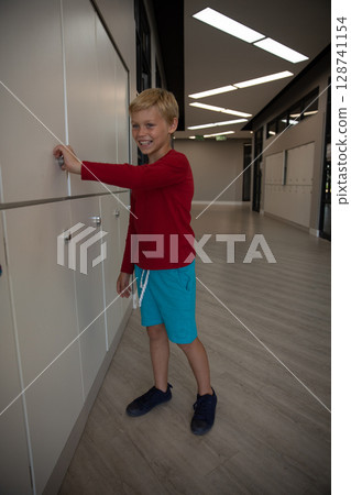 Young boy standing in modern indoor corridor turning white locker handle, smiling in red shirt Young boy standing in modern indoor corridor turning white locker handle, smiling in red shirt 128741154