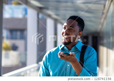 Young African American man using smartphone with backpack on covered outdoor walkway, smiling Young African American man using smartphone with backpack on covered outdoor walkway, smiling 128741156