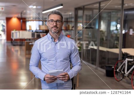 Man holding small spiral notepad in center of open-plan office, featuring books and bicycle 128741164